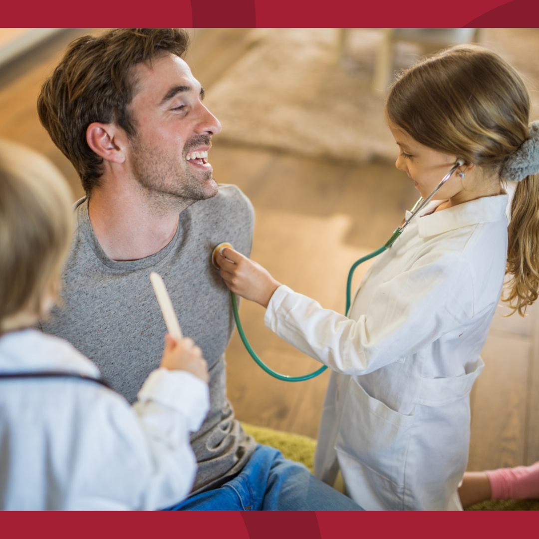 A man with two young children who are dressed like doctors. His daughter is pretending to listen to his heart with a stethescope.